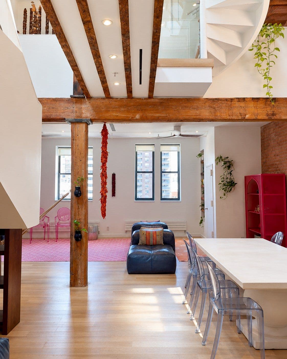 An open-plan living and dining room viewed from the kitchen end, with a long white table and clear chairs at right, a dark sofa in the center, and a pink tiled seating area at left. Exposed beams and a post cross the room beneath a mezzanine and curved staircase; daylight enters through three rear windows.