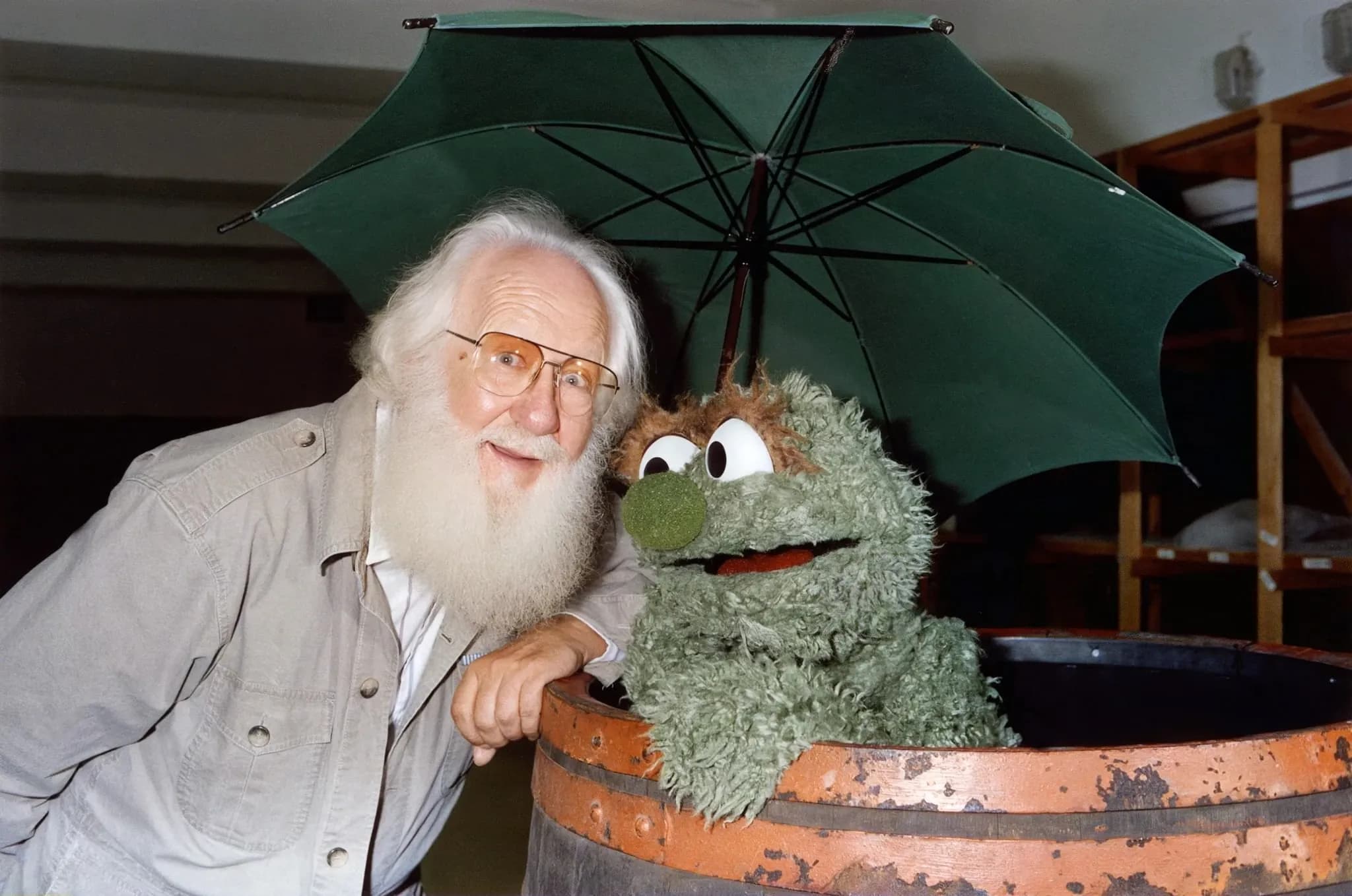 Kermit Love, white-bearded in glasses, leaning beside the Oscar the Grouch puppet in a barrel under a green umbrella