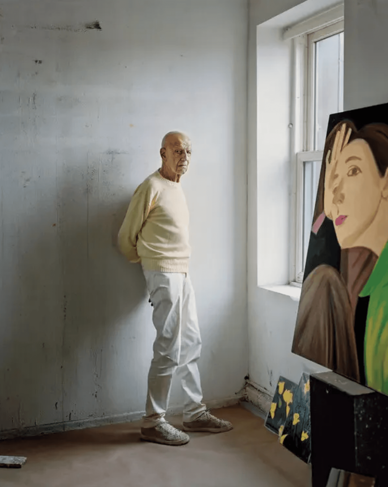 Alex Katz standing against a bare studio wall beside a window, with one of his large painted portraits in the foreground