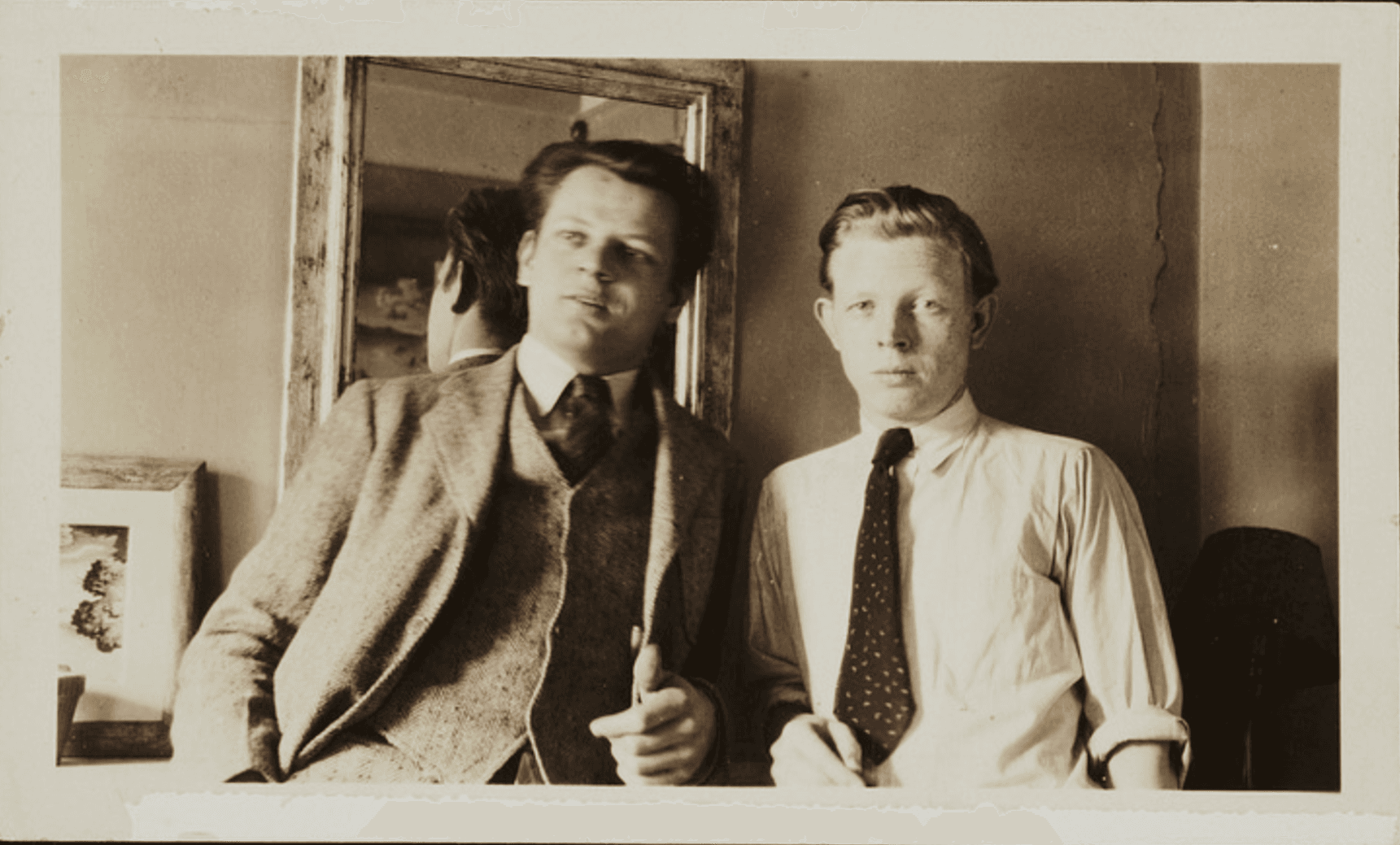 Sepia-toned photograph of two men posing side by side indoors. One wears a suit and leans against a surface; the other wears a light shirt with rolled-up sleeves and a dark tie. A mirror behind them reflects part of the first man's head.