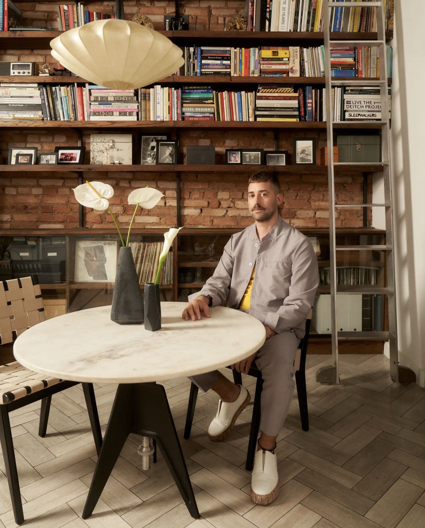 Gabriel Yuri sits at a round pale table in front of extensive built-in shelving with red brick backing. The shelves hold books, photographs, and objects; a cream pendant light hangs above the table. A dark vase with white flowers sits on the table, and a woven chair is visible on the left.