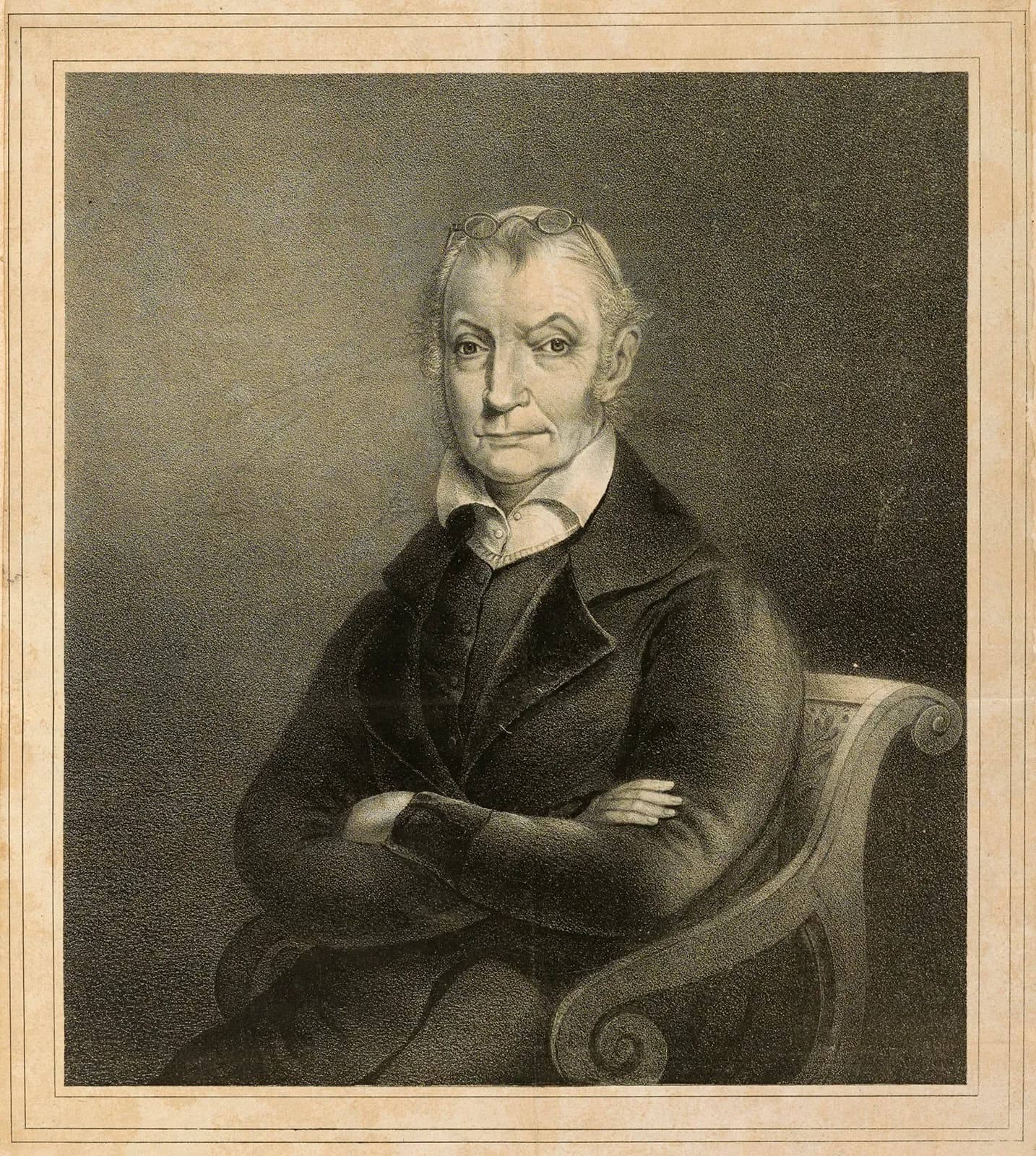 Monochrome portrait of an older man seated in a chair with his arms crossed, wearing a dark coat and glasses perched on his head.