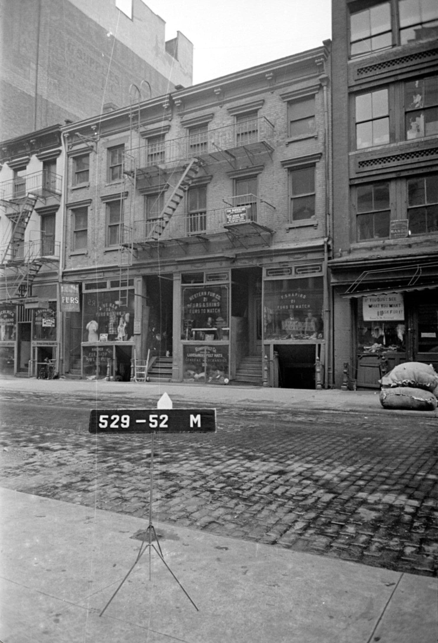 Black and white photograph of a brick building facade taken from street level. The building has multiple stories with regularly spaced windows and fire escapes on the front. Ground floor storefronts with display windows line the street, with a taller building visible behind on the left. A measurement marker labeled 529-52 M stands on the brick sidewalk in the foreground.