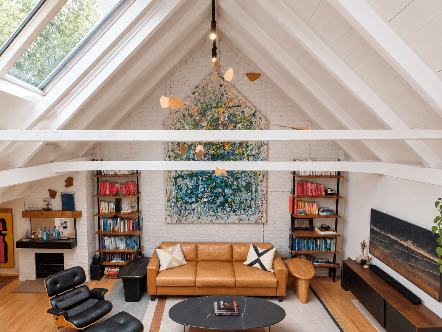 Bright living room with a vaulted white ceiling and skylight, a large colorful painting above a tan sofa, bookshelves on either side, and a black lounge chair and coffee table in the foreground.