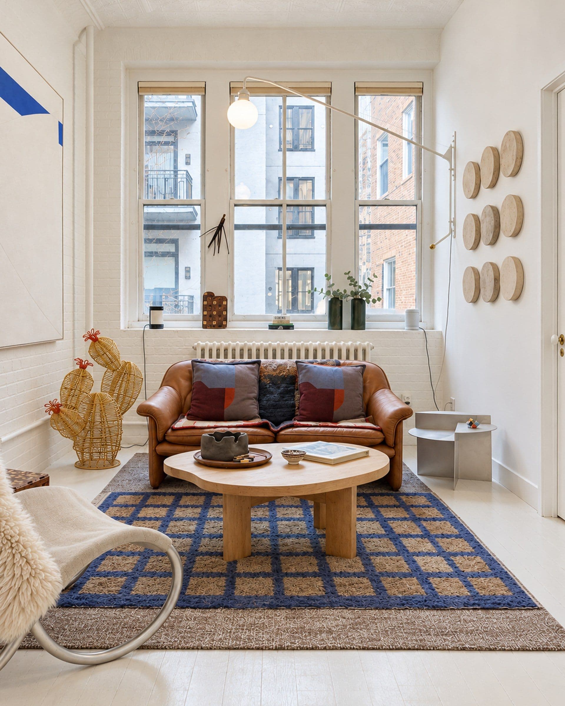 A living room seen from across a checked blue and tan rug toward three tall windows that look onto neighbouring buildings; a tan leather sofa with patterned cushions sits below the centre window with a white radiator behind it, and a round light-toned coffee table stands on the rug in front. The left wall is painted white brick hung with a large pale canvas marked with a blue shape, the right wall carries nine round discs in three rows and a long swing-arm lamp reaching over the sofa, and the floor is painted white boards.