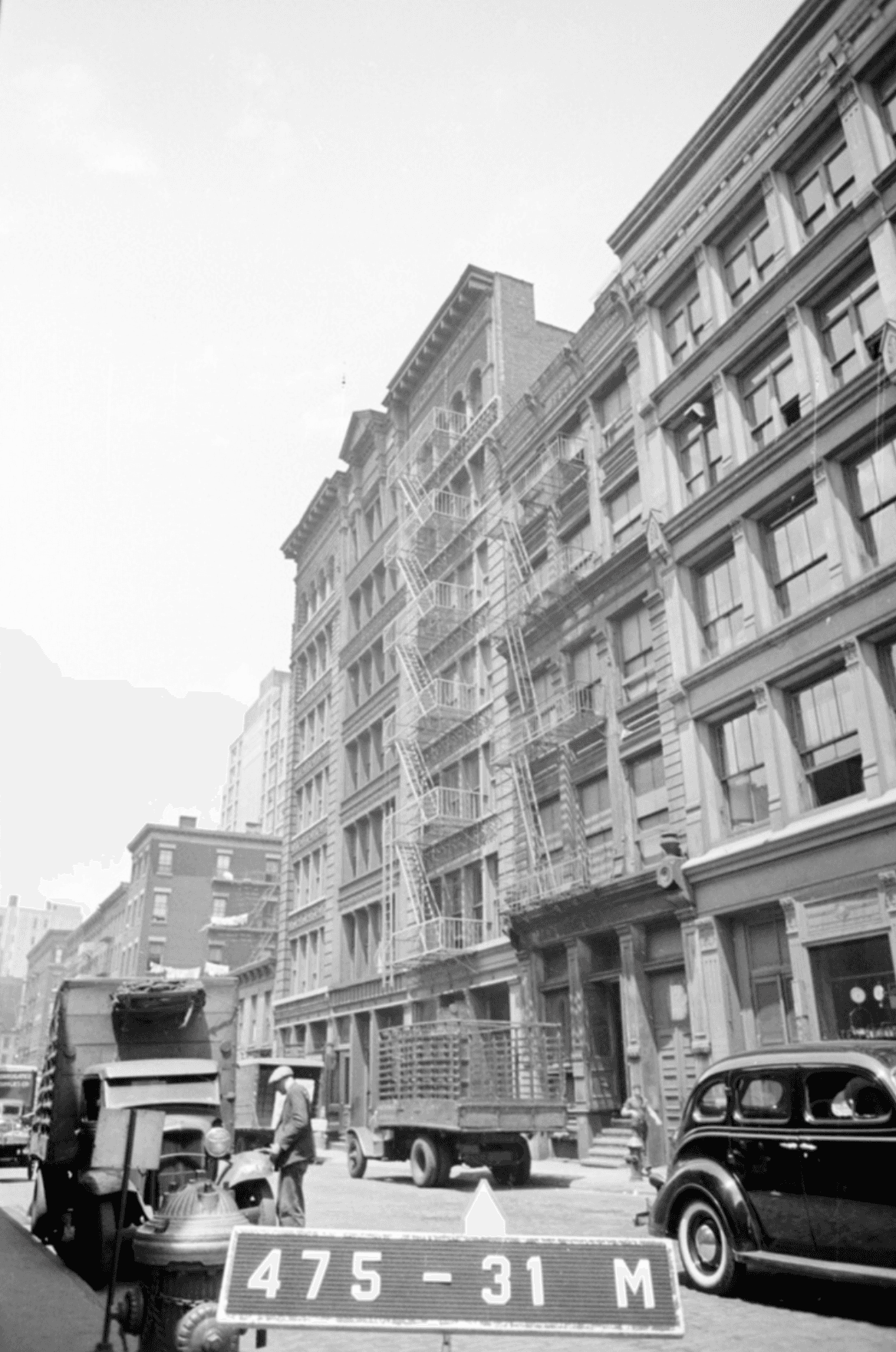 A black and white photo of an urban street with tall buildings, many fire escapes, several trucks, a classic car, and a man near a fire hydrant in the foreground.