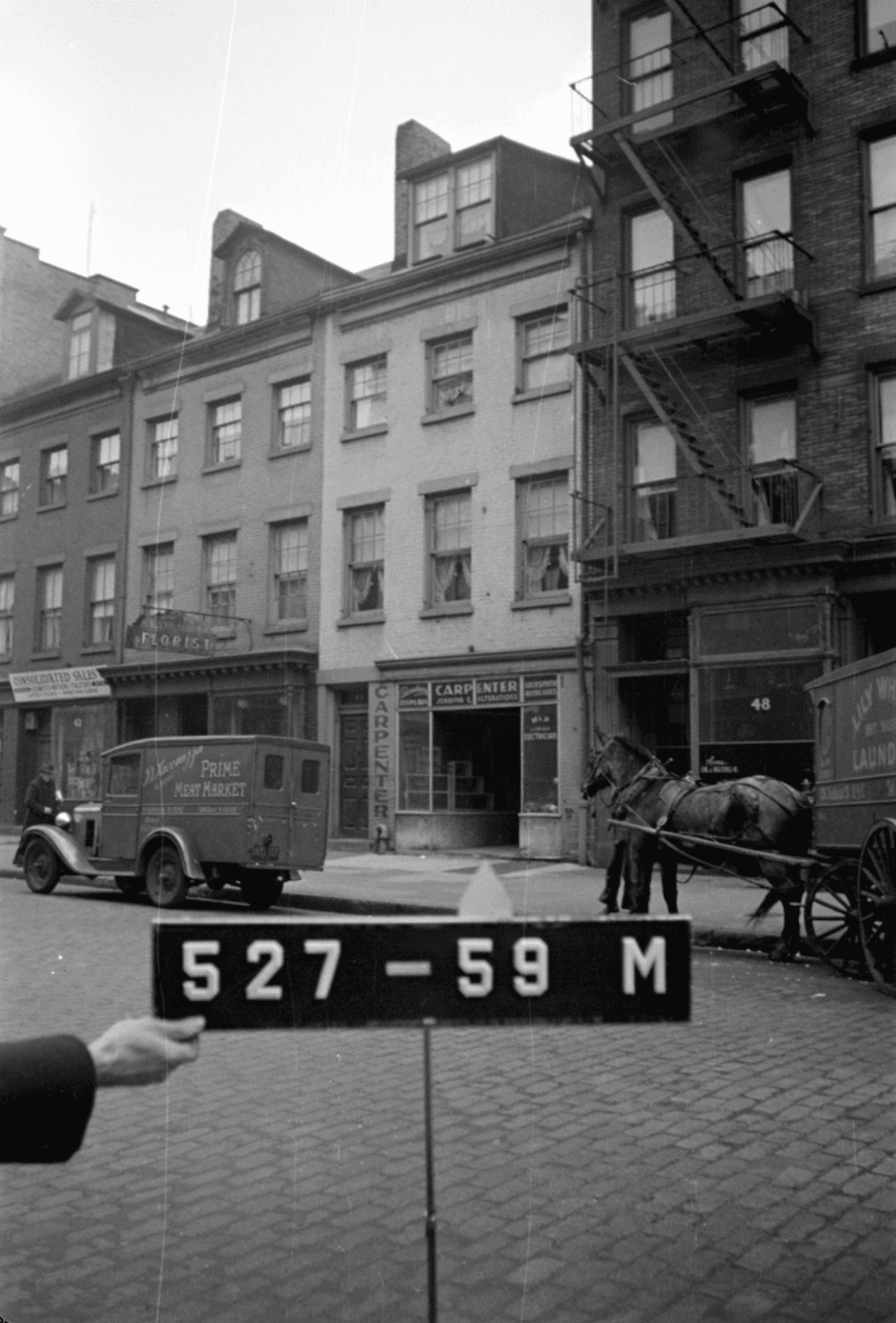 Black-and-white street scene with storefronts, a parked delivery truck, a horse-drawn wagon, and a person holding a large numbered sign in the foreground.