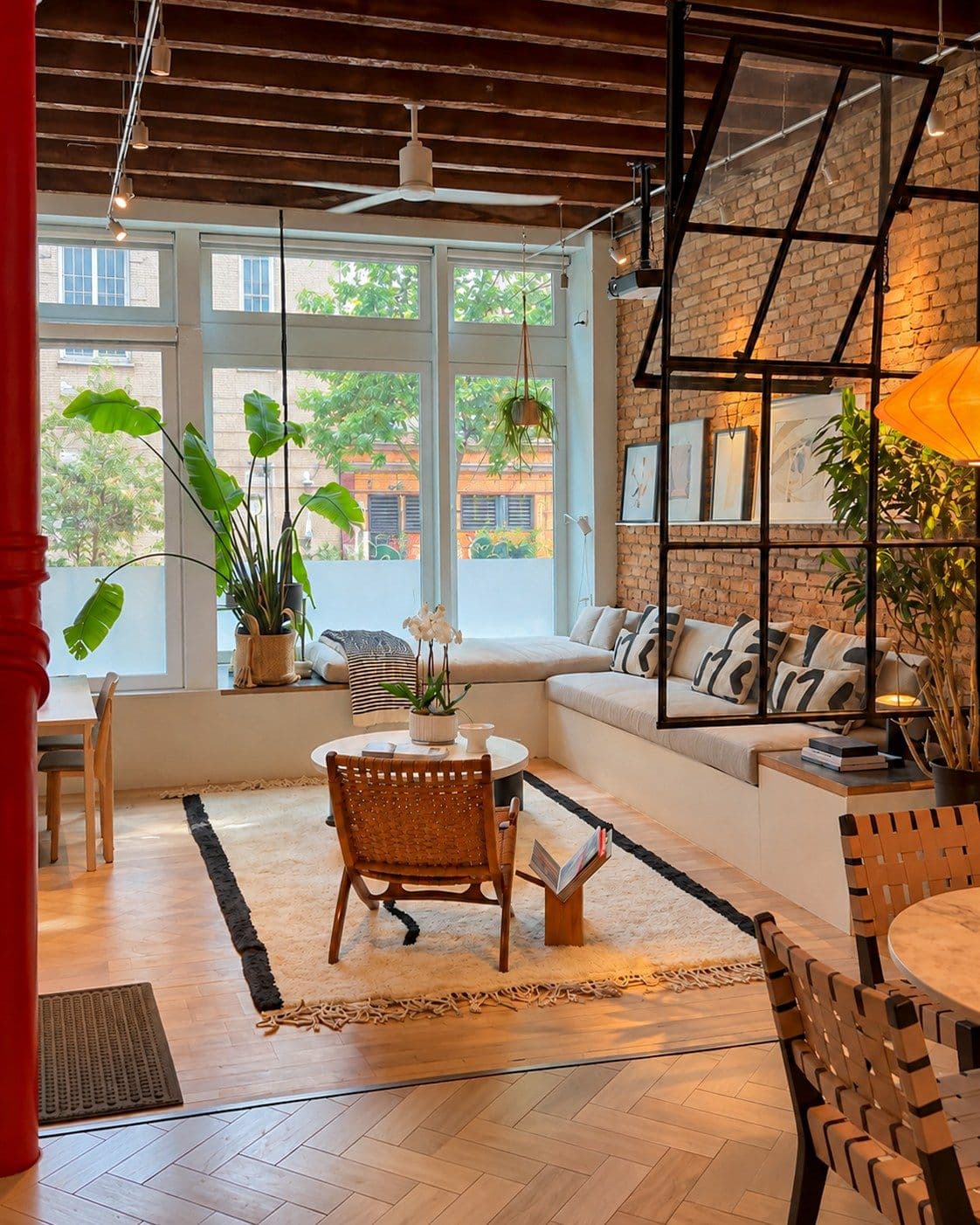 A loft-style living room with exposed wooden beams and brick wall on the right. Large windows on the far wall overlook buildings and green foliage outside. A cream-colored built-in seating bench with cushions runs along the brick wall, divided by a black metal grid partition holding potted plants. In the foreground, a woven chair faces a small round white table. Wooden chairs with woven seats sit on the right. Potted plants in various sizes are positioned throughout the room near the windows and along the brick wall. Light comes from the windows on the far wall.
