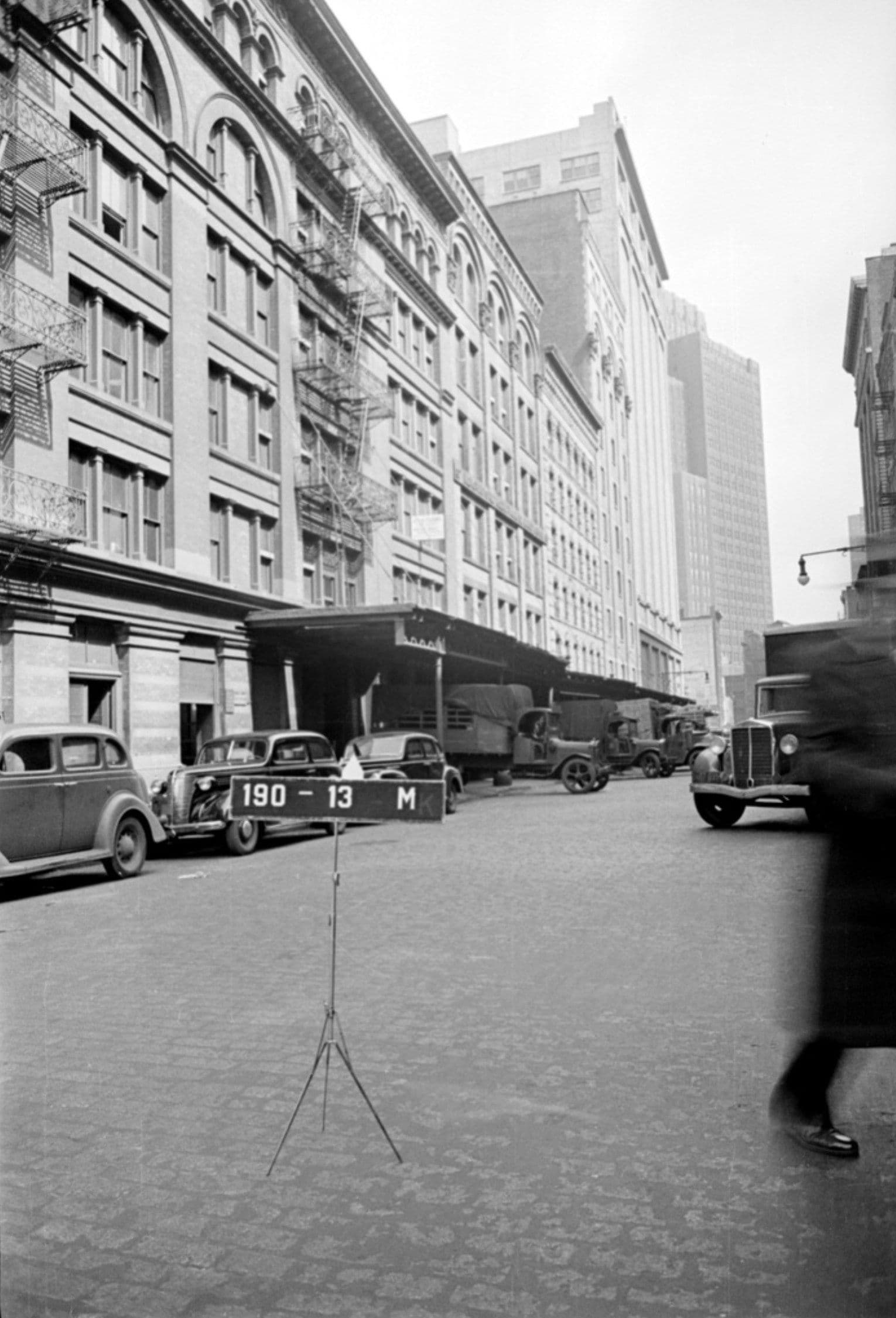 A black and white street photograph looking down a wide roadway lined with tall arched-window buildings fitted with fire escapes on the left. Cars and covered trucks are parked along the curb and beneath a loading canopy, a small sign reading 190-13 M stands on a tripod in the foreground, and a blurred pedestrian crosses on the right.