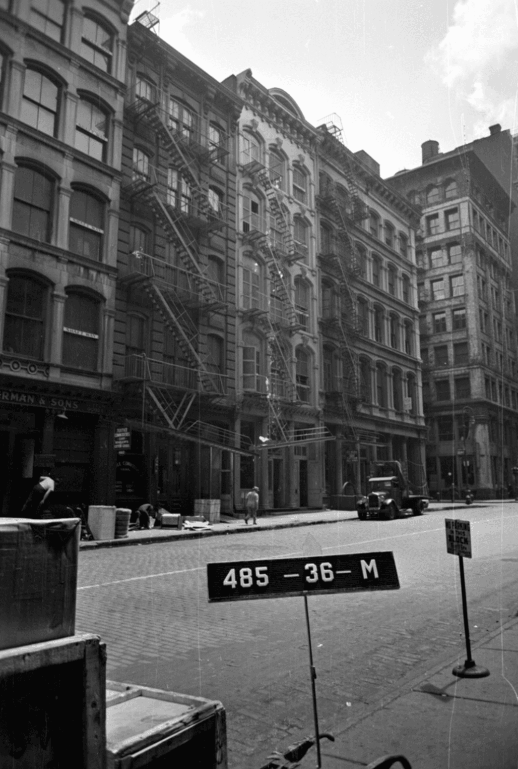 Black-and-white archival street photograph of cast-iron loft buildings with fire escapes, an old truck and a tax lot sign